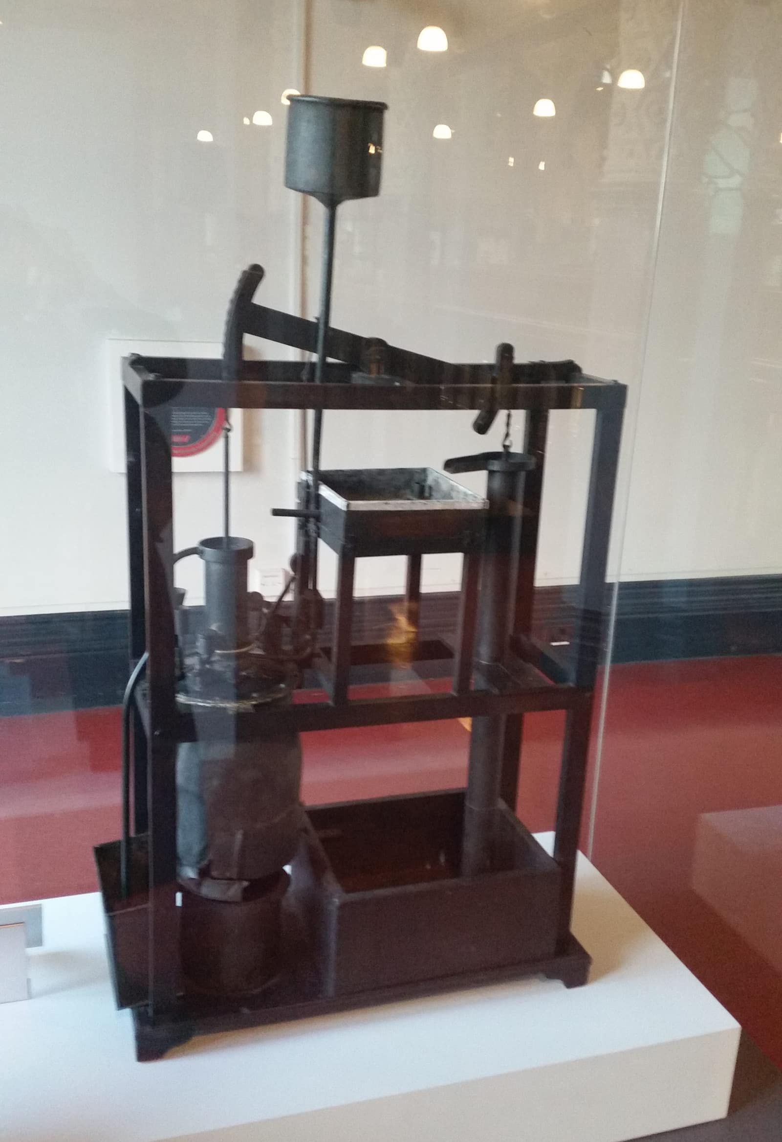 Dans une vitrine, une petite maquette de machine à vapeur, de la taille d'un enfant, montée dans un bâti ouvert de bois sombre. À sa base, à gauche, une chaudière ronde sur un foyer ; au-dessus, un cylindre vertical ; en haut, un balancier de bois qui oscille sur un pivot, une chaîne à chaque bout, l'une vers le piston du cylindre, l'autre vers une tige de pompe à droite. Une citerne est posée sur une tablette au milieu et un grand entonnoir de métal gris s'élève au-dessus du bâti.