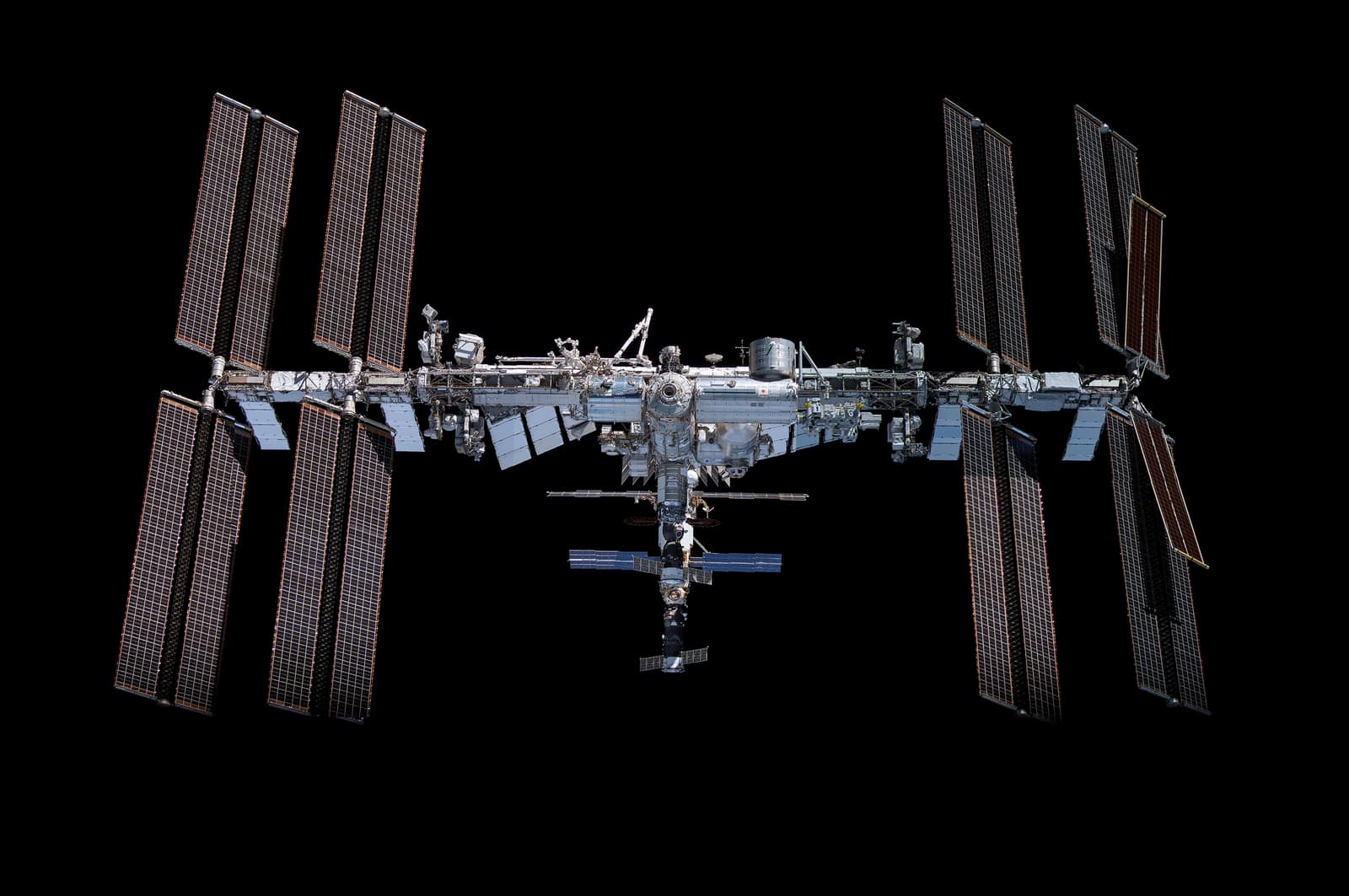 La Station spatiale internationale vue en entier sur le noir de l'espace : une longue poutre horizontale porte à chaque extrémité quatre grandes ailes de panneaux solaires brun cuivré, avec au-dessous des radiateurs clairs ; au centre, un groupe de modules cylindriques blancs et un laboratoire japonais arrondi, et une file verticale de modules pend vers le bas, avec de plus petits panneaux solaires gris-bleu et un vaisseau amarré à son extrémité inférieure.
