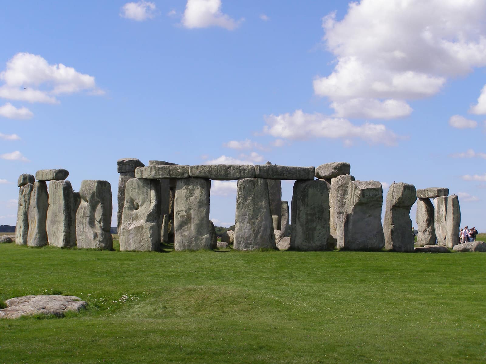 Les pierres dressées de Stonehenge sur une herbe verte rase, sous un ciel bleu vif parsemé de nuages blancs : un cercle de montants massifs, gris et striés de lichen, dont plusieurs sont encore reliés par de longs linteaux horizontaux, avec des vides là où des pierres sont tombées ; au premier plan gisent des pierres plus petites et un bloc abattu, et un groupe de visiteurs se tient au bord droit.