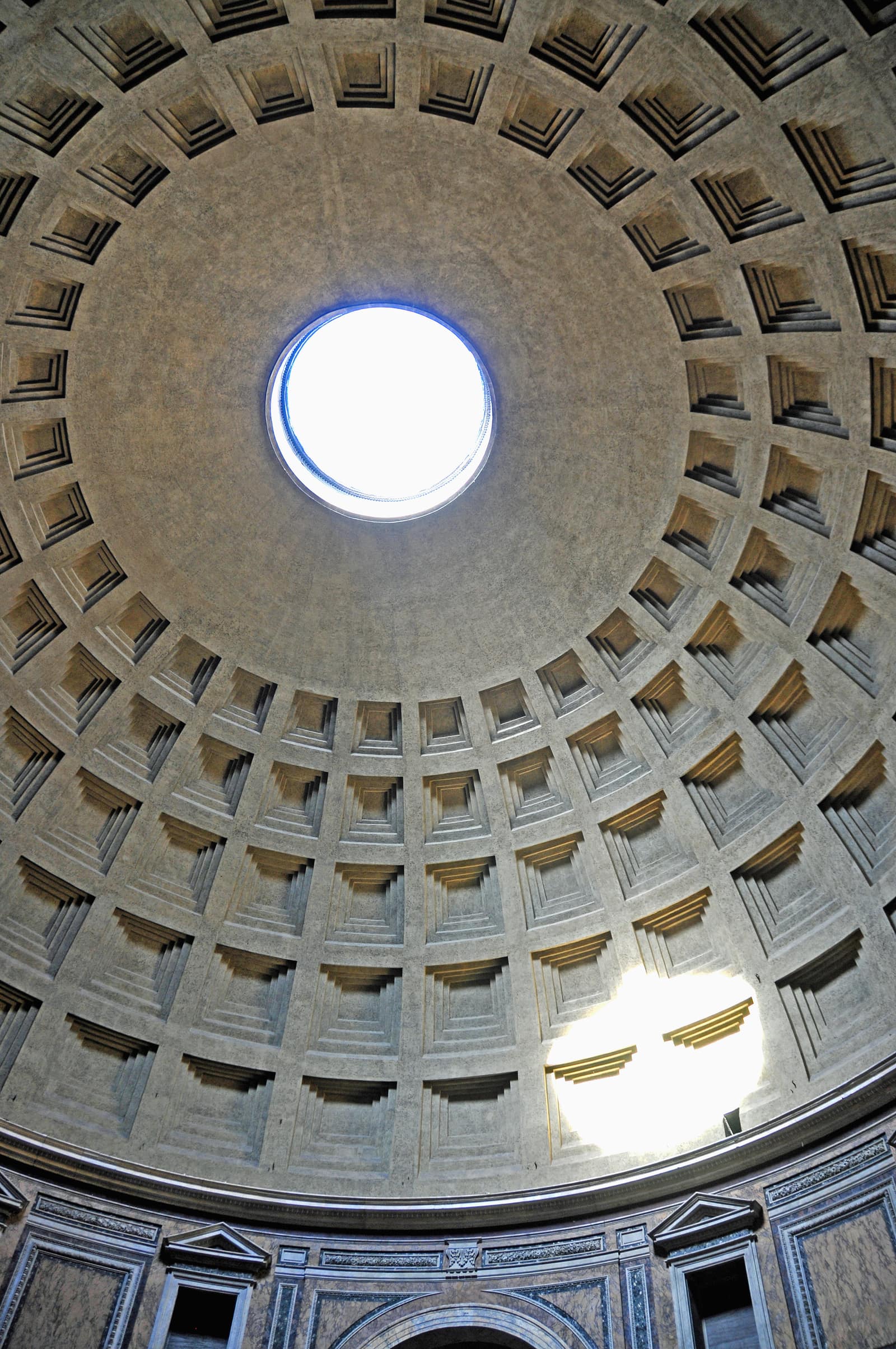 Vue prise en regardant droit vers le haut dans la coupole du Panthéon à Rome : des rangées de caissons carrés, chacun creusé de plusieurs ressauts, rapetissent en montant vers une ouverture ronde au sommet, d'un blanc éclatant de ciel. La surface de béton est grise et marbrée. Une tache de soleil éclaire les caissons en bas à droite, et une corniche de marbre ponctuée de niches court au bas de l'image.