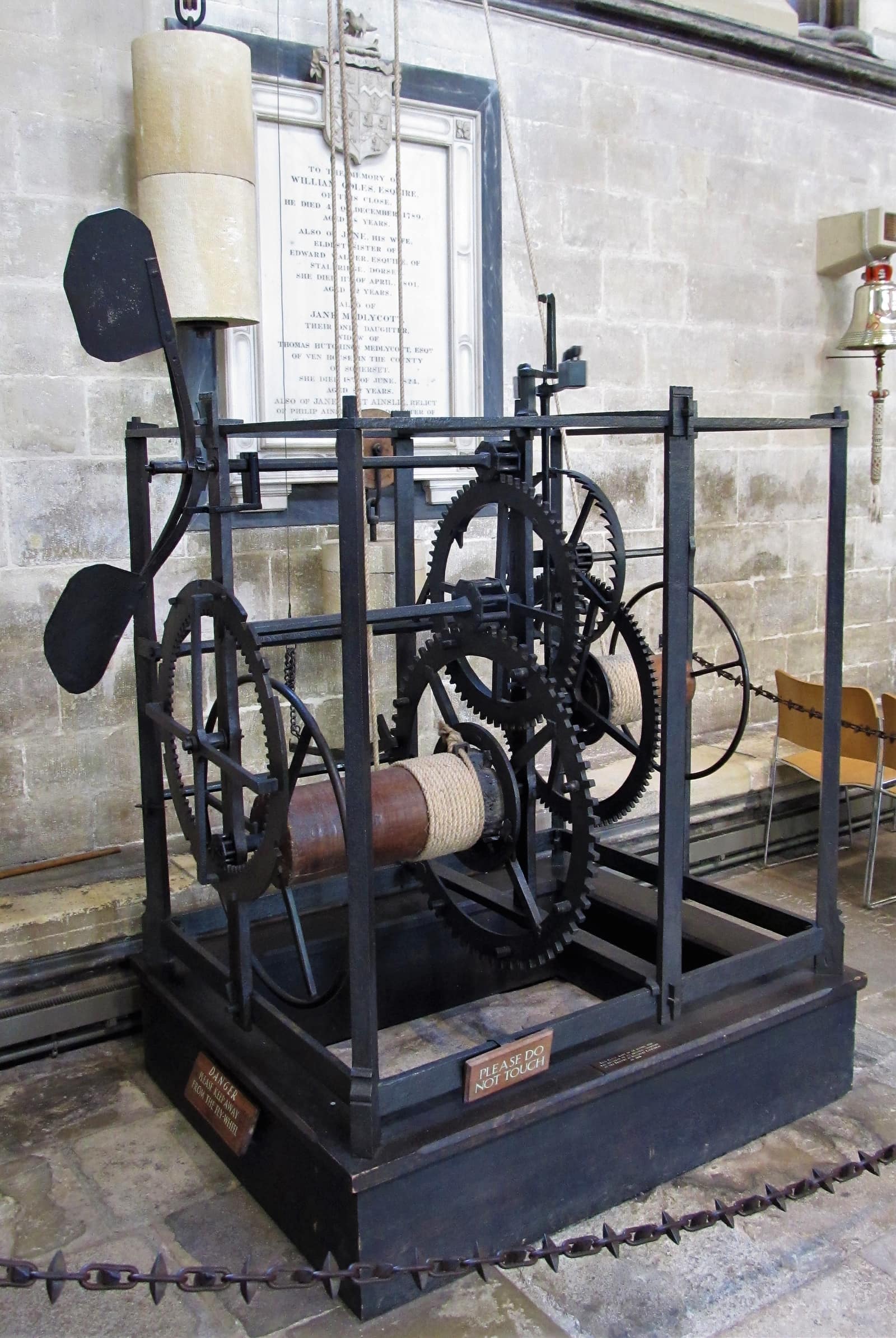 Une cage ouverte de fer forgé noir, de la taille d'un grand coffre, posée sur un socle bas et sombre contre le mur de pierre claire d'un bas-côté de cathédrale. À l'intérieur, de grandes roues dentées de fer et deux tambours de bois enroulés d'une épaisse corde claire ; à gauche, un long bras mobile se termine par deux palettes noires et plates. Des cordes montent des tambours vers de lourds poids de pierre suspendus au-dessus ; derrière l'horloge, une plaque funéraire de marbre ; un petit écriteau porte la mention Please do not touch.
