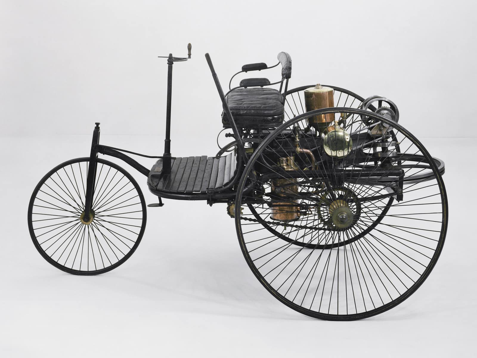 Vue de profil, sur un fond blanc uni, d'une voiture légère à trois roues. Une seule roue à rayons à l'avant, de la taille d'une roue de bicyclette, est dirigée par une tige verticale munie d'une petite manivelle ; deux roues bien plus grandes, aux rayons fins, portent l'arrière. Sur un plancher de lattes de bois repose une banquette noire rembourrée ; derrière et dessous se trouvent le moteur et ses organes, un cylindre de laiton, un globe de verre, des récipients de cuivre et, à l'arrière, un volant horizontal.