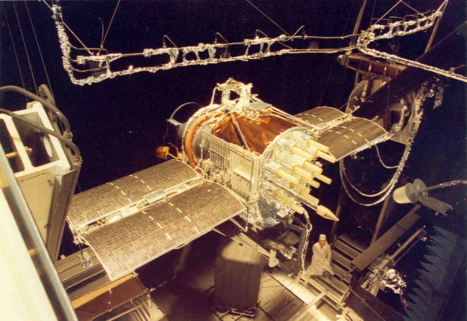 Photographie en couleur d'un satellite dans une immense chambre d'essai obscure. L'engin, enveloppé de feuilles d'or et d'argent, a un corps en forme de caisson, un faisceau de courtes tiges crème pointées vers l'avant et deux longs panneaux solaires plats déployés comme des ailes de part et d'autre. Des câbles et un cadre de tubes gainés de feuille métallique pendent au-dessus ; tout en bas, une petite silhouette en combinaison et coiffe blanches de salle propre se tient près du support.