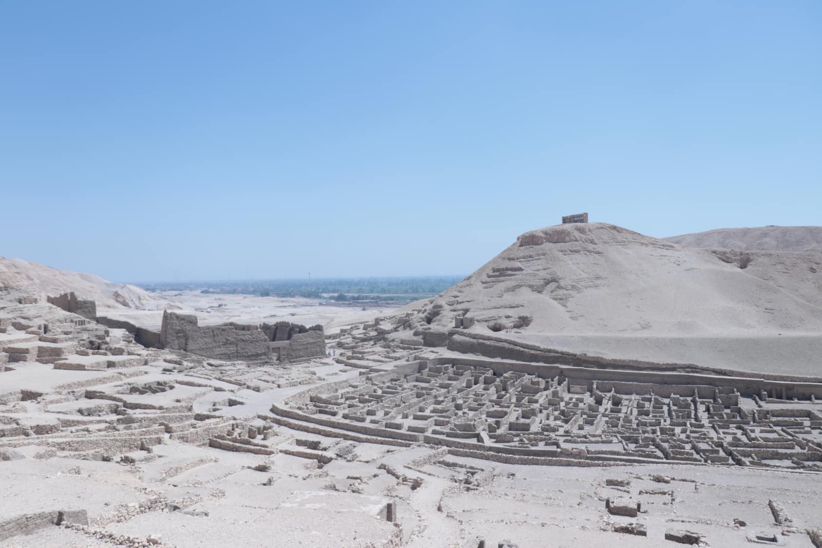 Les murets de pierre claire d'un village en ruine, serrés en un quadrillage de petites pièces et de ruelles étroites, au pied d'une colline de sable nue sous un ciel bleu sans nuage ; à gauche se dresse un mur d'enceinte plus haut, en ruine, et au-delà des collines désertiques s'étire, dans la brume, une bande de champs verts de la vallée du Nil.