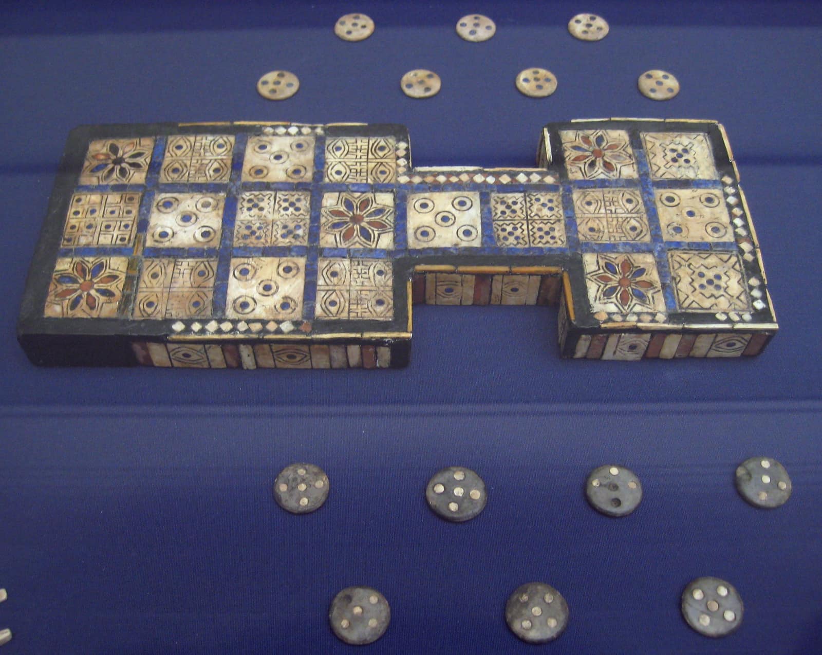 Un petit plateau de bois dans une vitrine, sur un tissu bleu sombre, formé de deux blocs de cases reliés par un étroit pont de deux cases : ses vingt cases de coquillage sont incrustées de rouge et de bleu, cinq d'entre elles portent des rosettes à huit pétales, d'autres des cercles de points ou des motifs en forme d'œil, le tout serti de fines baguettes bleues ; au-dessus et au-dessous, deux rangées de pions ronds, sept clairs à cinq points sombres et sept sombres à cinq points blancs.