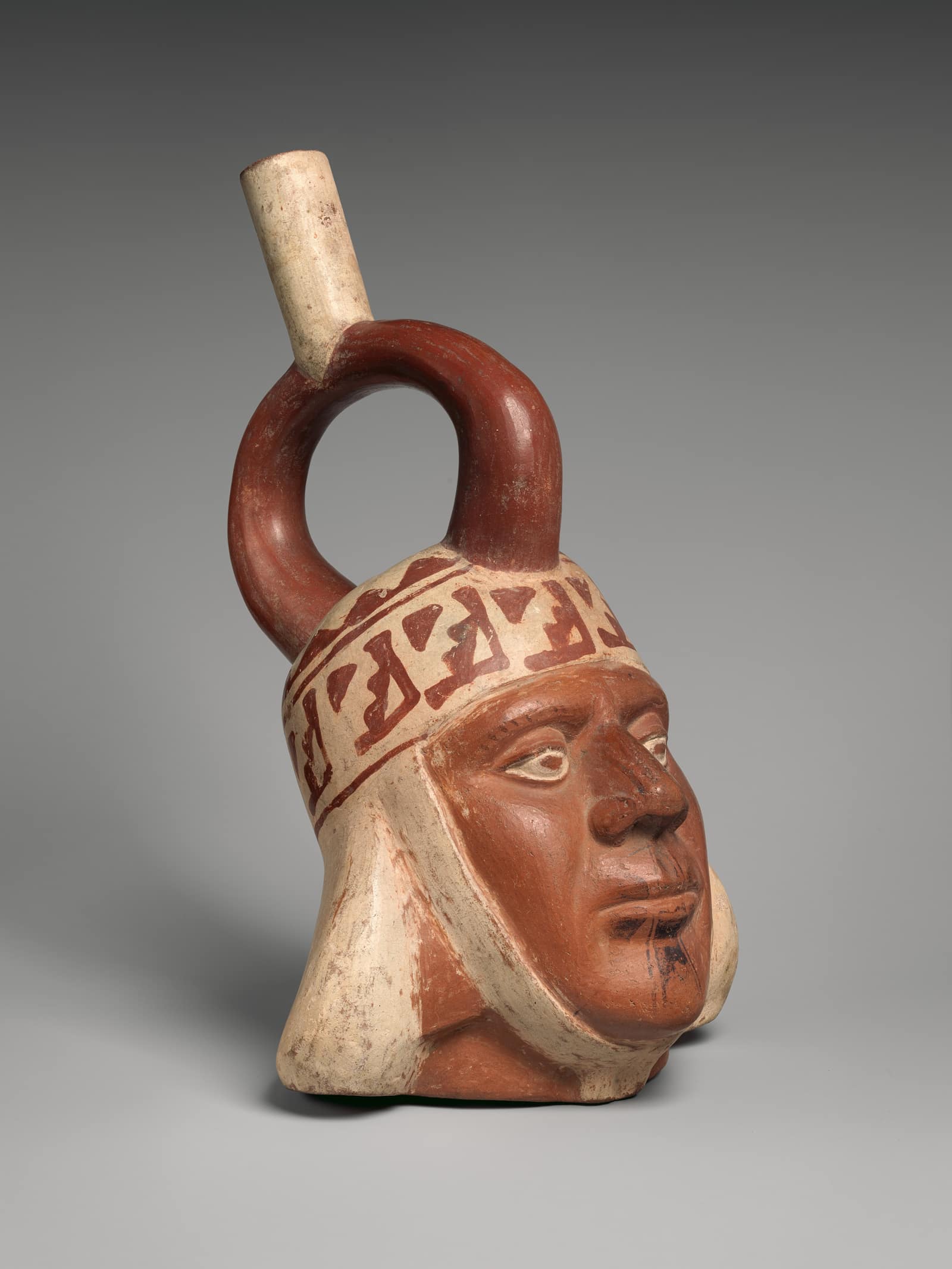 Une bouteille de céramique en forme de tête d'homme, vue de trois quarts sur fond gris : visage brun rouge au nez affirmé, lèvres pleines entrouvertes, yeux cernés de blanc et de noir ; une coiffe de tissu crème ornée d'une bande de motifs géométriques rouge sombre encadre le visage, et une anse en étrier terminée par un goulot crème s'élève au sommet. De fins traits noirs sont peints sous la lèvre inférieure.
