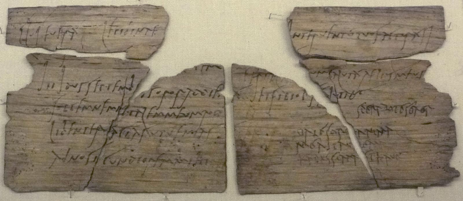 Quatre fragments pâles et veinés d'une mince tablette de bois, posés sur un tissu beige en deux moitiés séparées par un étroit intervalle. Des lignes d'encre sombre et passée, en cursive romaine, courent sur le bois ; dans le bas de la moitié droite, quelques lignes plus courtes sont tracées d'une autre main, plus serrée.