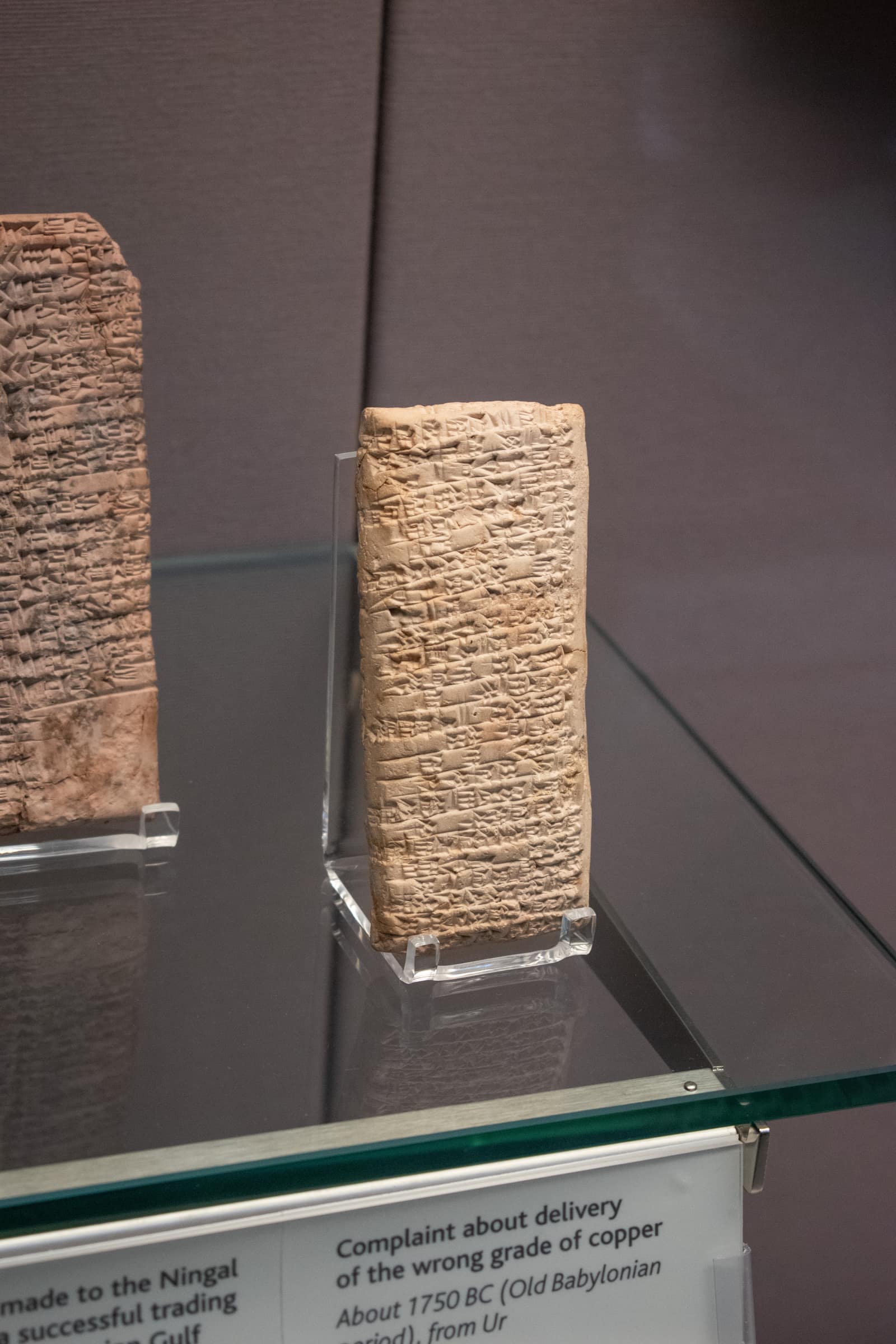 Une petite tablette d'argile dressée, couleur de sable pâle, couverte d'un bord à l'autre de lignes serrées de signes cunéiformes en forme de coins, posée sur un support transparent dans une vitrine de musée. Le coin d'une autre tablette inscrite apparaît à gauche, et une étiquette au premier plan commence par : Complaint about delivery of the wrong grade of copper.