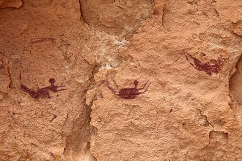 Une paroi de grès orangé et rugueux sur laquelle trois petites figures humaines sont peintes en rouge sombre. Chacune est allongée à l'horizontale, tête ronde, bras tendus vers l'avant et jambes repliées vers l'arrière, comme en train de nager. Entre elles, la surface de la roche s'est fendue et détachée par plaques pâles.