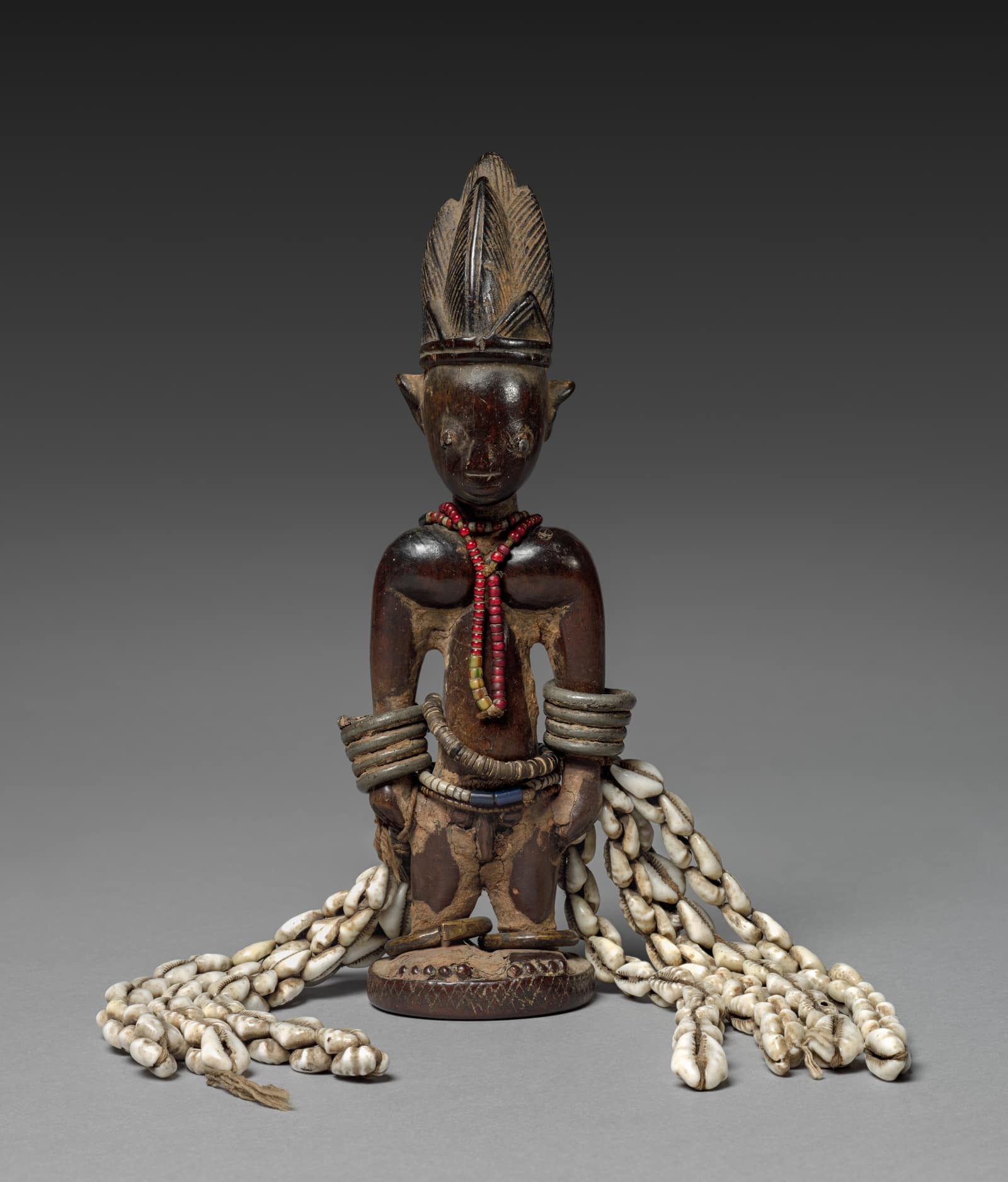 Une petite figure de bois debout sur un socle rond, photographiée sur fond gris : un adulte à la haute coiffure en crête, au visage étroit dont les yeux, le nez et la bouche sont usés jusqu'à être presque lisses. Le corps sombre et poli montre des plages claires là où la surface s'est effacée. Il porte un collier de perles rouges, des anneaux de métal empilés sur les deux bras et une ceinture de perles, et de longs rangs de cauris blancs tombent de sa taille jusqu'au sol, de chaque côté.